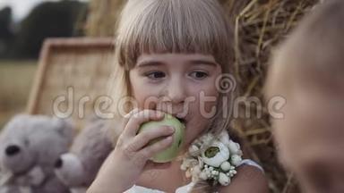 小女孩吃一个绿色的苹果。 酷酷的小男孩和小女孩坐在干草堆下面
